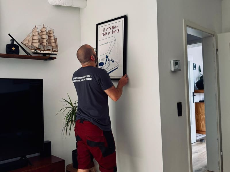 Cem richtet im Wohnzimmer einen gerahmten Kunstdruck an der Wand aus.